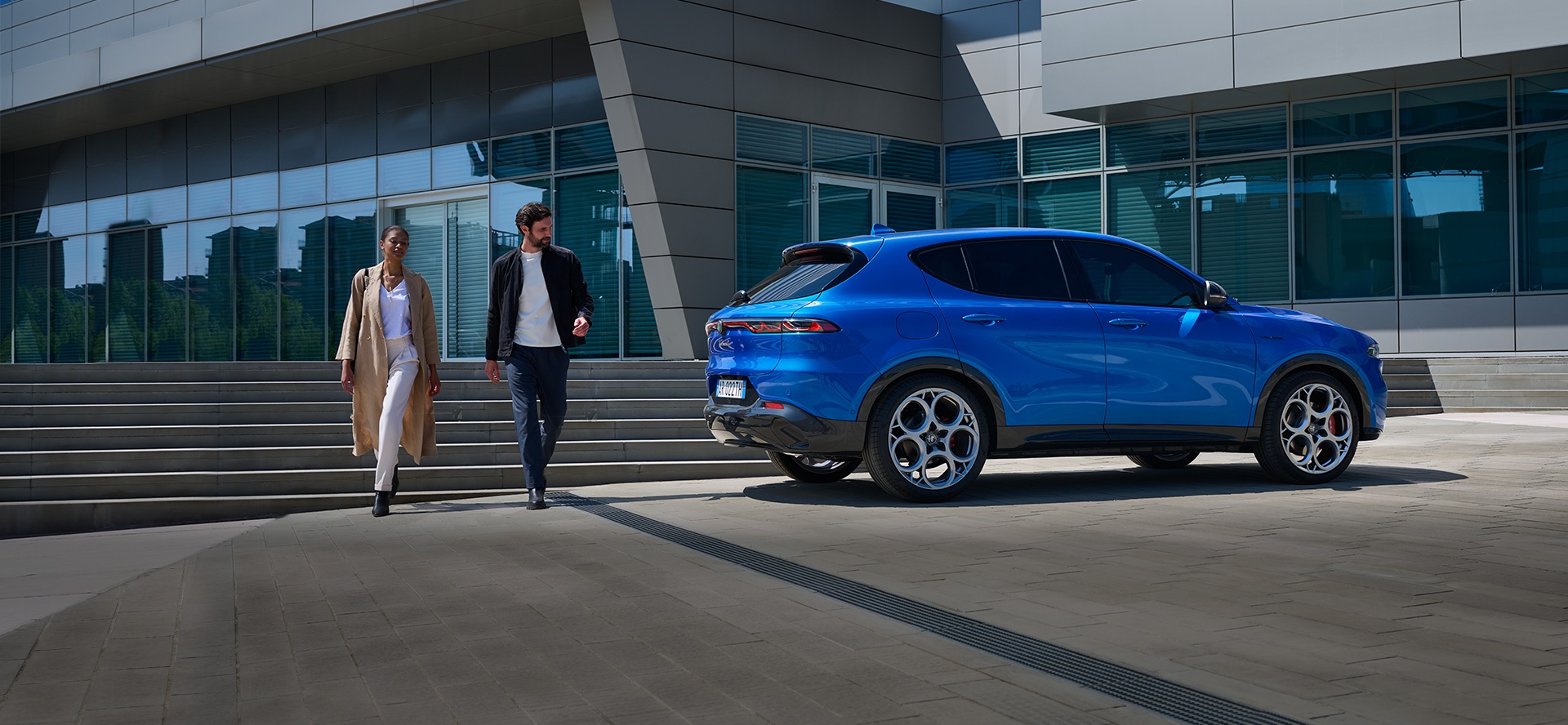 Kobieta i meżczyzna na tle schodó do biurowca, obok błędkitne auto z marek oferowanych przez grupę Stellantis
