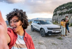 Kobieta wyciągająća rękę w kierunku obiektywu, w tle białe auto z resztą rodziny